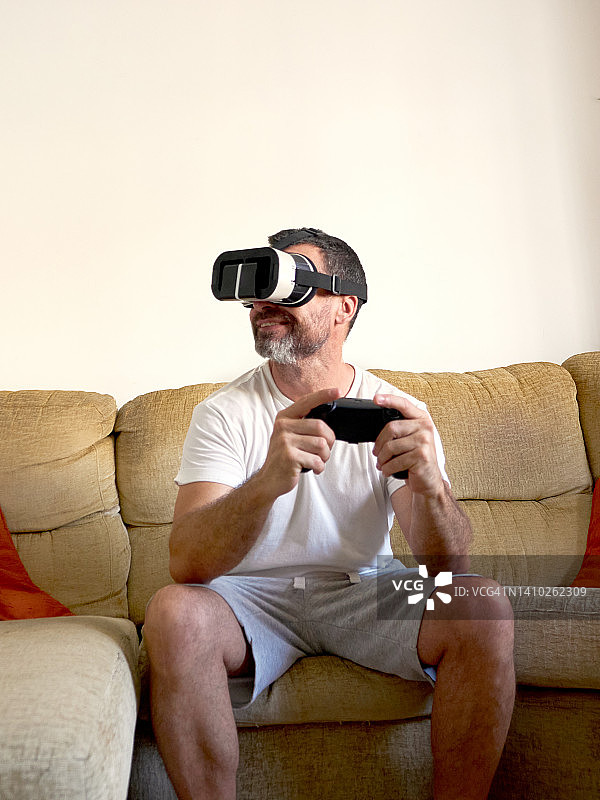 中年男子（40-45岁）留着胡须坐在沙发上，戴着虚拟现实眼镜用控制器玩电子游戏图片素材