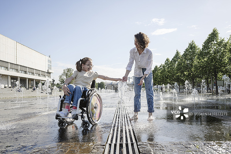 坐在轮椅上在公园里和母亲一起玩喷泉的女孩图片素材