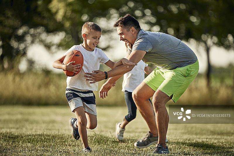 快乐的男人在运动场上追逐抱着橄榄球奔跑的儿子图片素材