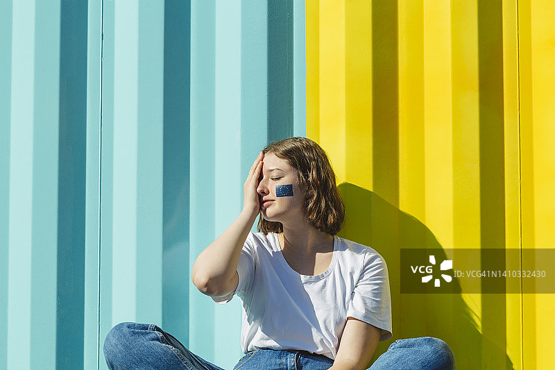 在蓝黄色墙前，面部涂有欧盟标志的少女遮住眼睛图片素材
