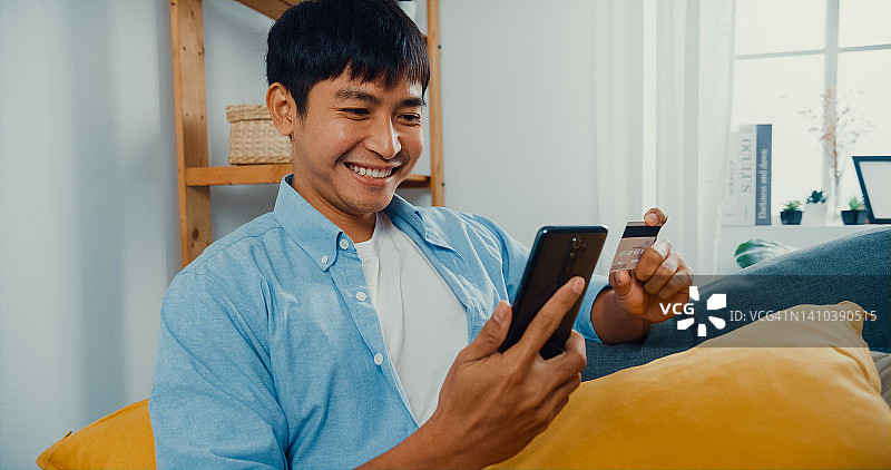 在家中客厅的沙发上使用手机和信用卡进行在线购物的年轻男人图片素材