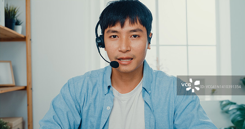 年轻男人在家中客厅的工作桌旁，戴着耳机，看着摄像头，用笔记本电脑与客户进行视频通话图片素材
