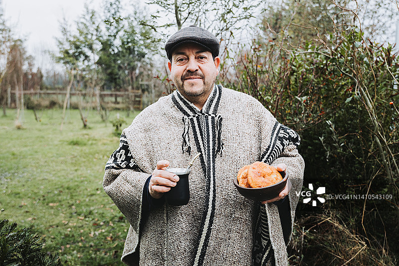 身穿灰色羊毛披风、手持马黛茶和盛着索派皮亚斯黏土碗的马普切族传统男子图片素材