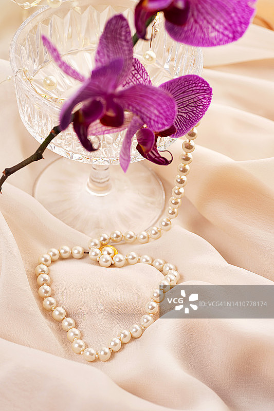 心形珍珠饰品、鸡尾酒杯和米色薄纱上的紫色兰花，微距拍摄图片素材