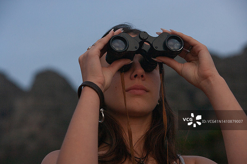 一位美丽的年轻女子专注于远方天空中的某物，一个留着黑色长发的少女正在训练她的眼睛，并用双筒望远镜放大远处物体图片素材