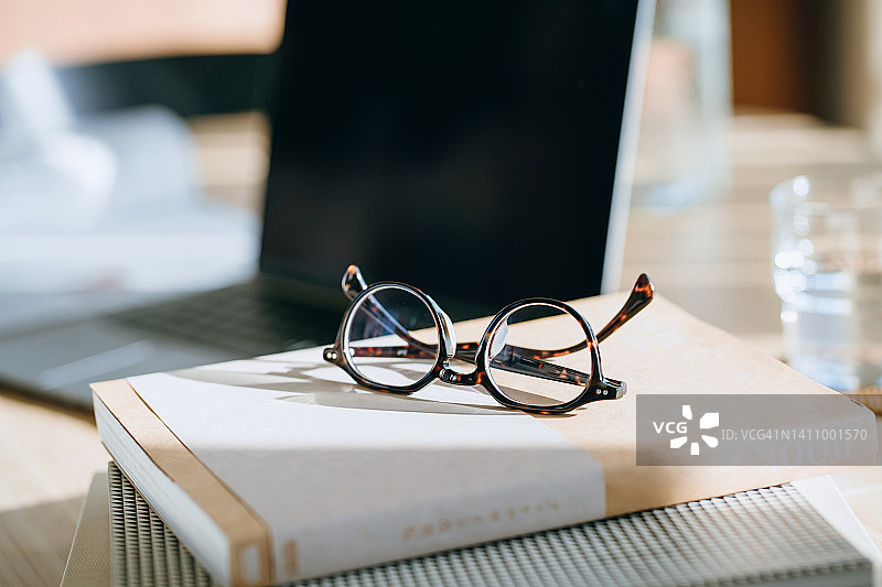 工作区桌子上眼镜特写，背景有书籍、文件和笔记本电脑，自然阳光。居家办公概念图片素材