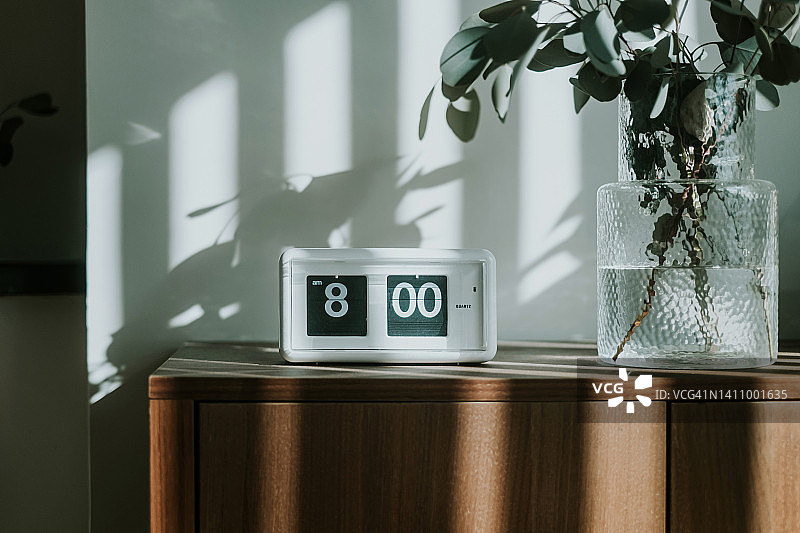 白色数字翻页时钟显示八点，位于客厅新鲜桉树旁的棕色木柜上，阳光透过窗户照耀，清新美丽的早晨：全新的一天，新的开始，新的能量，新的机遇图片素材