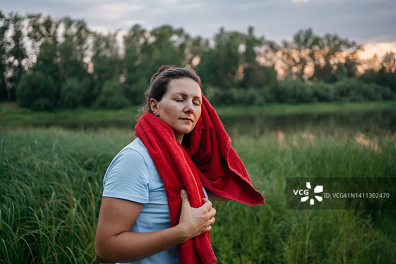 锻炼后疲惫的女人用毛巾擦脸，背景是湖图片素材