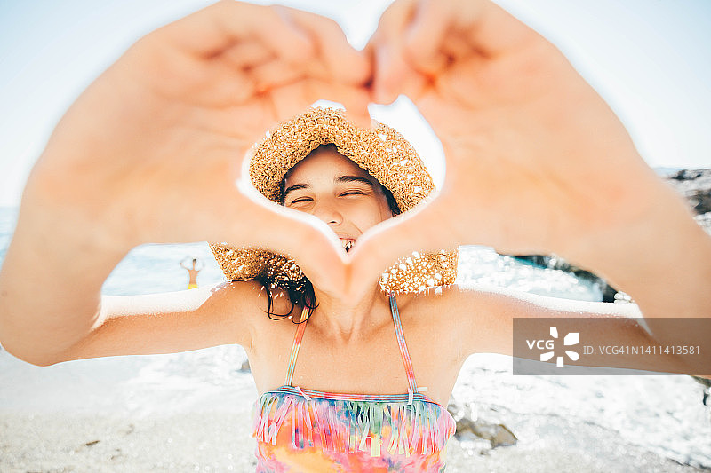 在沙滩上，戴着草帽，用手比心的欢笑少女肖像照图片素材