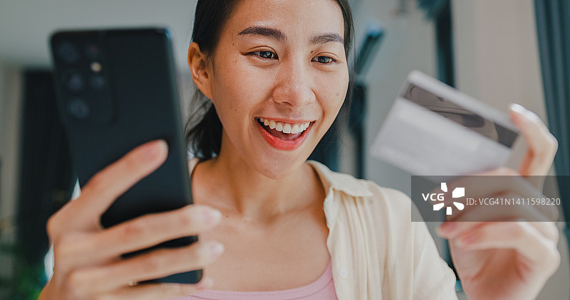 亚洲年轻女孩坐在桌前，手持智能手机和信用卡，在客厅里为网上购物支付成功感到高兴图片素材