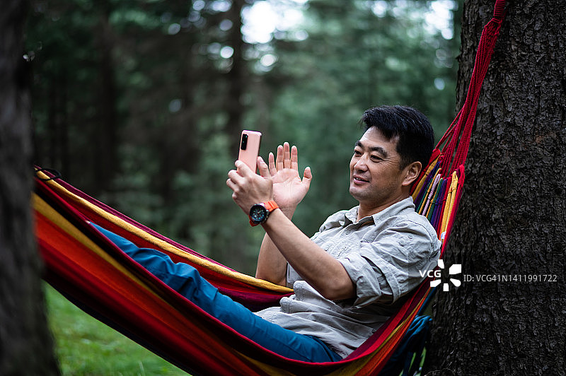 在森林里躺在吊床上使用智能手机的亚洲中年男人图片素材
