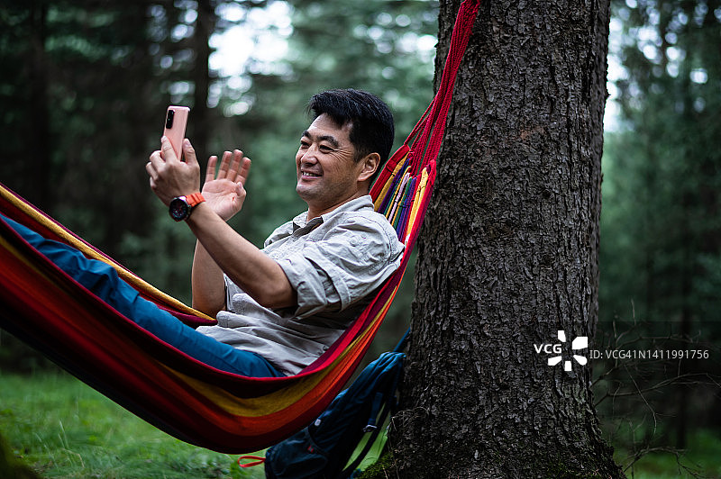 在森林里躺在吊床上使用智能手机的亚洲中年男人图片素材