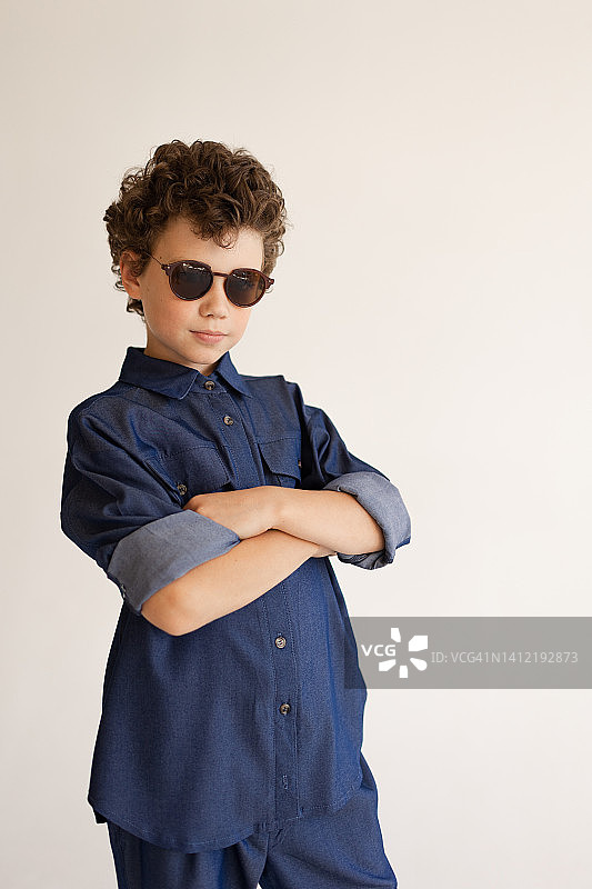 卷发男孩戴着太阳镜，身穿牛仔衬衫和牛仔裤在白色背景工作室的肖像图片素材