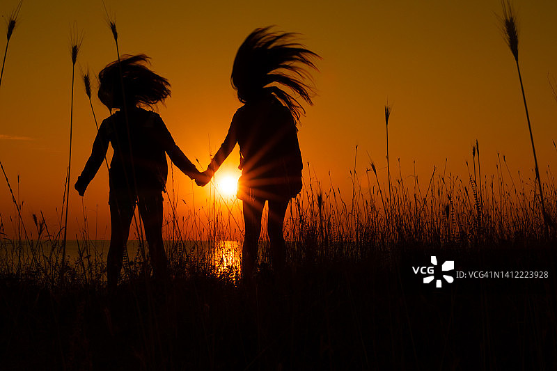 日出剪影：两名女子在悬崖顶上手牵手，吹着头发，欣赏着海边壮丽的景色图片素材