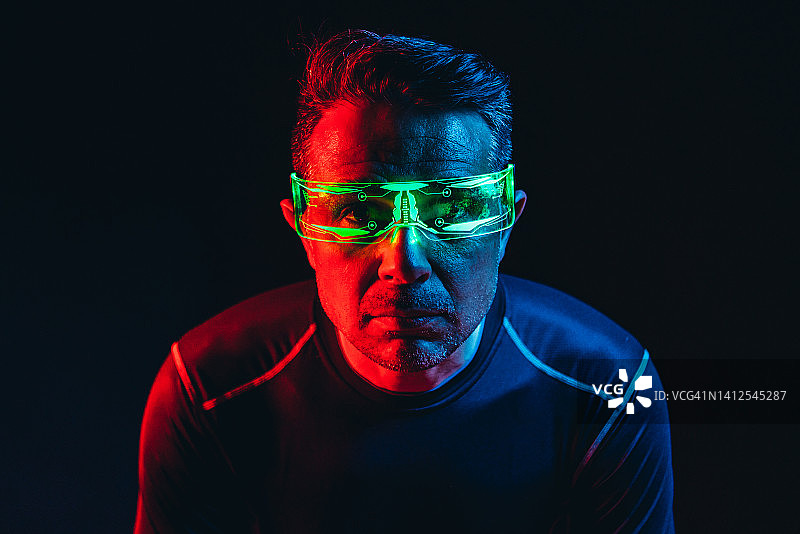 戴着绿色灯光电子眼镜、身穿蓝色紧身T恤的男人，在蓝红色环境中看向镜头，黑色背景。机器人、赛博格、未来和元宇宙概念图片素材