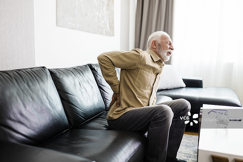 年长的男人感到不适，患有背痛，坐在家里的沙发上按摩酸痛的肌肉。老年人与健康问题概念图片素材