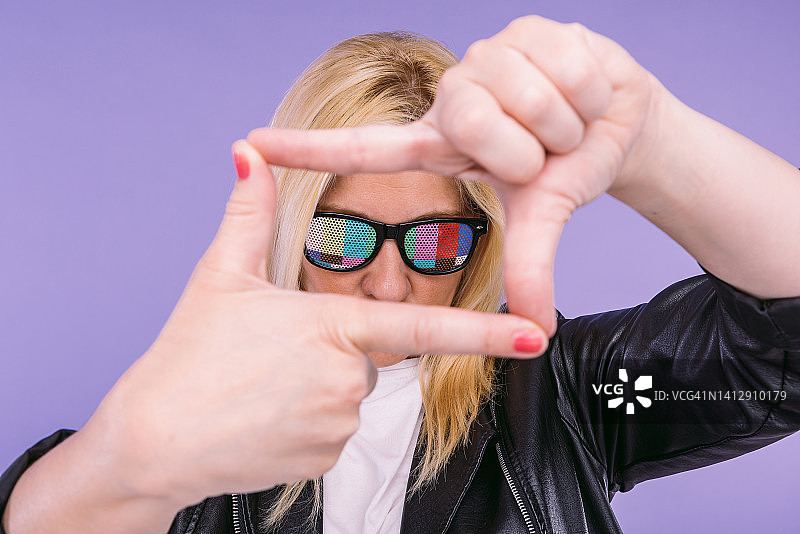 戴眼镜的金发女人手持电视测试卡，穿着黑色皮夹克，用手取景，背景为紫色。电视、广播、发射、电影和视听。图片素材