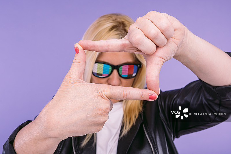 戴着眼镜的金发女人拿着电视测试卡，失焦，穿着黑色皮夹克，用手取景，在紫罗兰色背景上图片素材