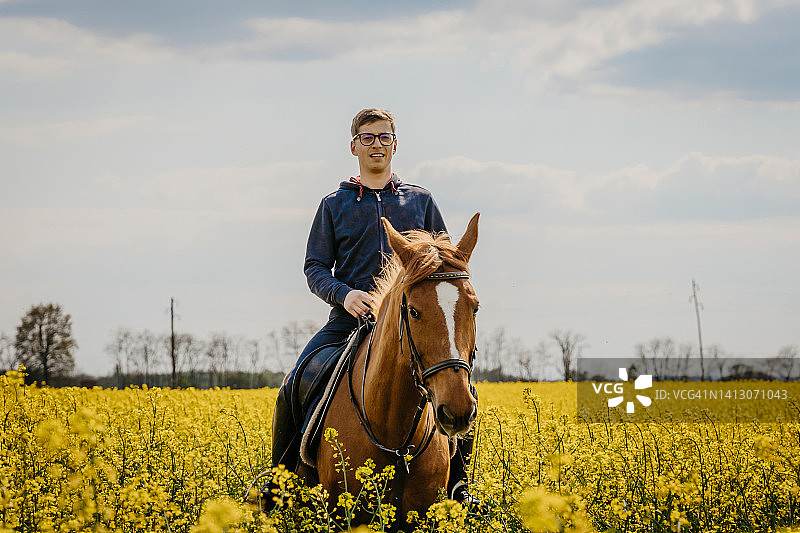 微笑的男人在多云的天空下骑马穿过油菜籽农场图片素材