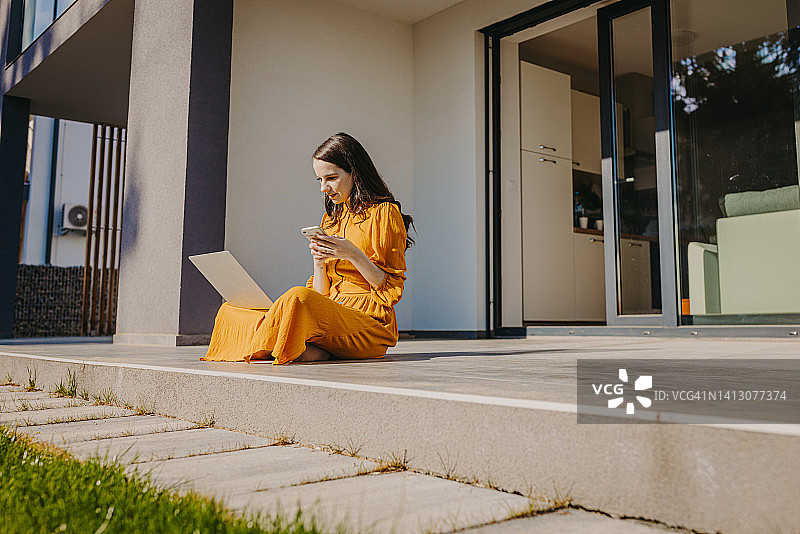阳光明媚的日子里，女人坐在门廊上用笔记本电脑和手机图片素材