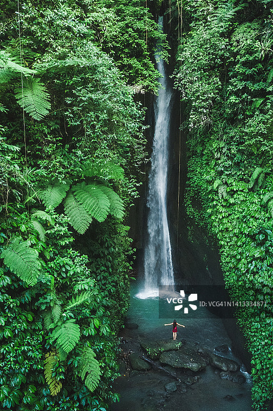 快乐的女人在巴厘岛美丽的勒克勒克瀑布附近举起双手。从后面鸟瞰图片素材
