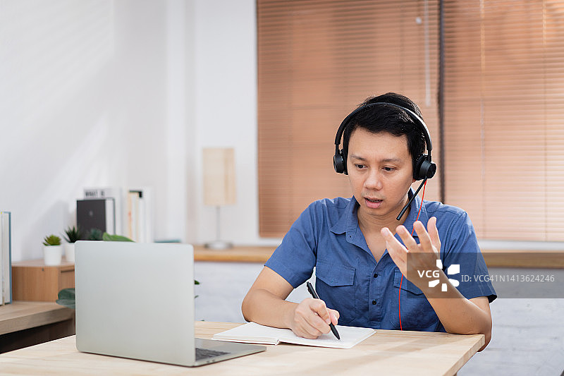 中年亚洲男性戴着耳机坐在家庭办公室里，使用电脑笔记本电脑为客户提供呼叫中心支持图片素材