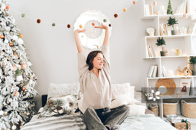 年轻漂亮女子坐在装饰有圣诞树和灯的房间的床上伸展身体，新年节日概念，正面视角图片素材