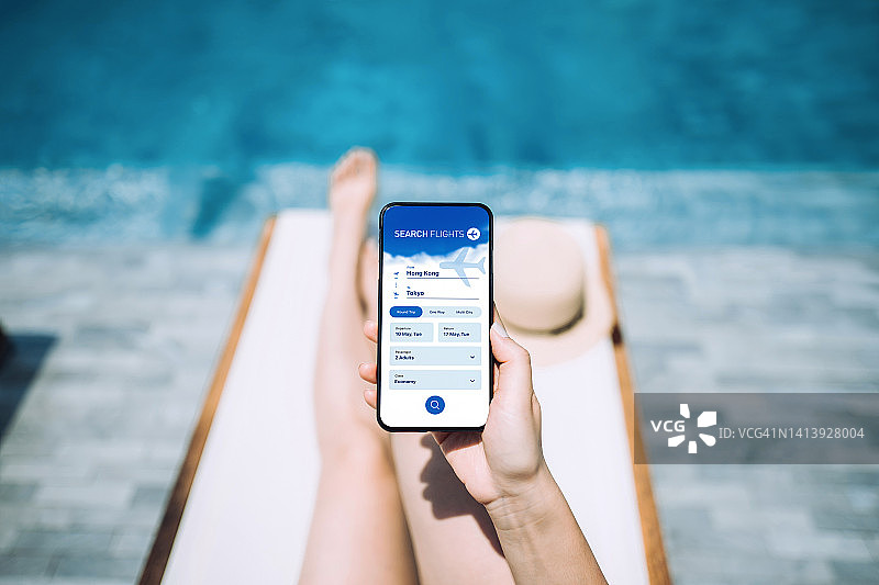 年轻女人在泳池边躺椅上享受阳光，同时使用智能手机特写：在航空公司网站上预订机票，进行旅行规划，在线预订假期，旅行和度假概念图片素材