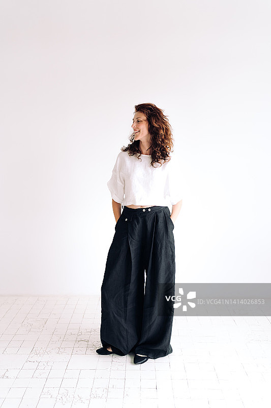靠在白色背景上，手放在裤子口袋里，笑容满面的棕色卷发年轻女子肖像。她穿着宽松的亚麻休闲服。自然美概念图片素材