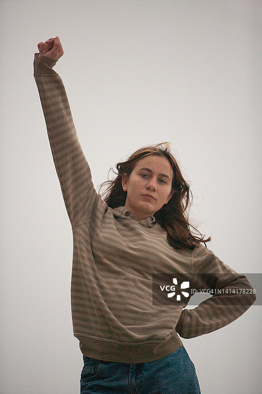 一位留着棕色长发和挑染刘海的美丽少女，穿着牛仔裤和条纹上衣，一只手放在臀部，另一只手臂高举图片素材