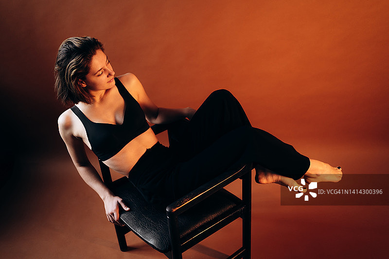 穿着黑色内衣和裤子的美丽女人坐在棕色墙前的黑色椅子上，留着短发，展现自然之美图片素材