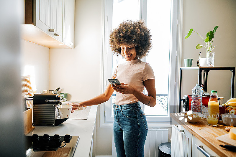 非裔美国卷发女子在家中厨房用咖啡机制作拿铁并使用智能手机图片素材