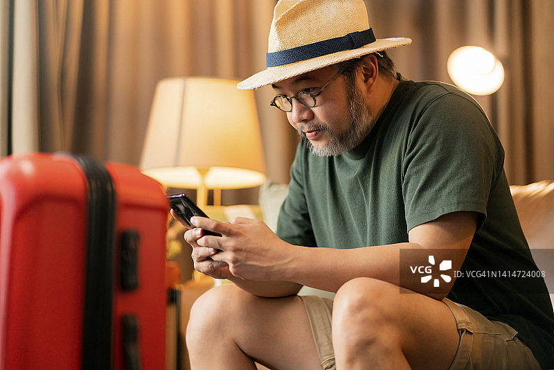 准备出国旅行，亚洲成年男子身穿休闲服装坐在沙发上，旁边放着行李旅行包，通过在线应用程序智能设备预订酒店房间和机票，东方旅行图片素材