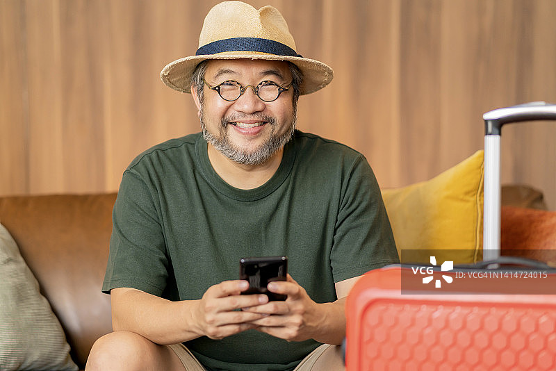 准备出国旅行，亚洲成年男子身穿休闲服装坐在沙发上，旁边放着行李旅行包，通过在线应用程序智能设备预订酒店房间和机票，东方旅行图片素材