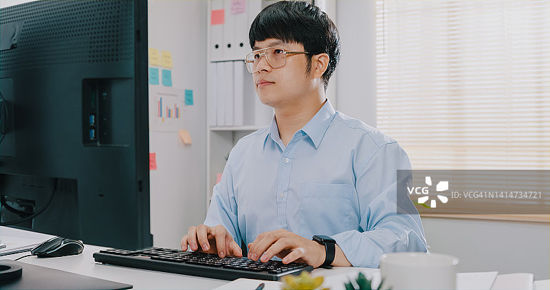 年轻的亚洲男商人身穿蓝色正装衬衫，坐在办公桌前，看着电脑打字，并在小型办公室检查报告文件，为会议报告做营销准备图片素材