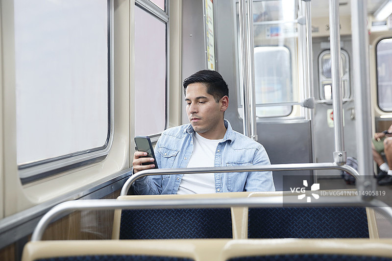 一位深色头发的拉丁裔千禧一代男人穿着商务休闲装坐在地铁上使用手机。图片素材
