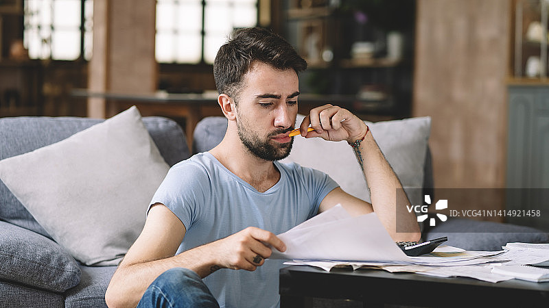 年轻男人坐在客厅沙发上，处理财务问题，应对经济和通货膨胀问题，支付账单，计算个人财务图片素材