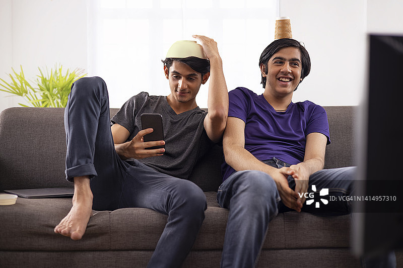 两个少年坐在沙发上，头顶碗和塑料杯，玩得很开心图片素材