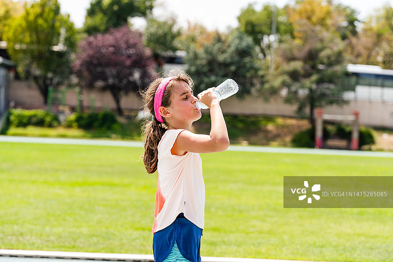 小女孩在田径俱乐部的跑道上休息喝水。女孩站着，从瓶子里大口喝水图片素材