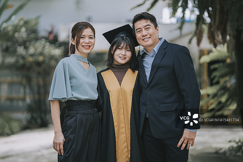 骄傲的亚洲父母和他们穿着毕业礼服的女儿在大学建筑外合影图片素材