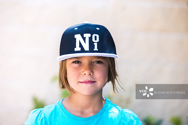 金发女孩戴着印有No. 1字样的黑白棒球帽，身穿绿色T恤，对着镜头微笑。儿童，肖像，漂亮，可爱，情感概念图片素材