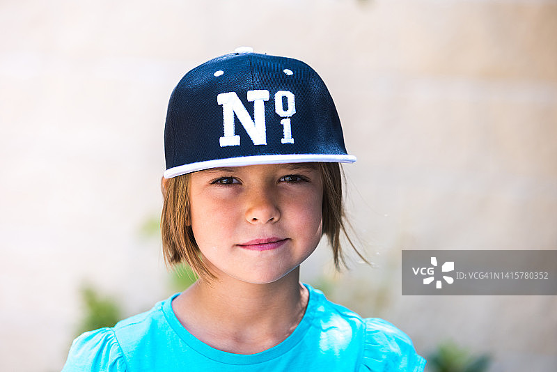 金发女孩戴着印有No. 1字样的黑白棒球帽，身穿绿色T恤，对着镜头微笑。儿童，肖像，漂亮，可爱，情感概念图片素材