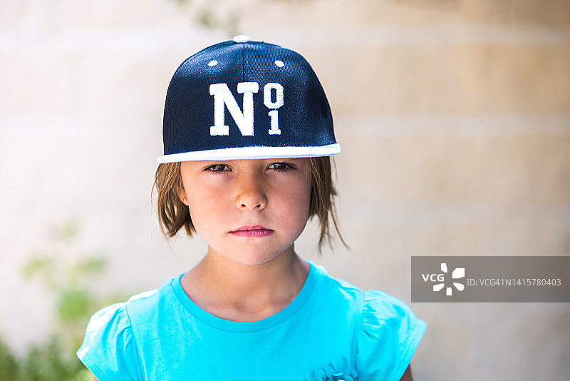 身穿绿色T恤、头戴黑白棒球帽的金发女孩，面容严肃，看向一侧。儿童时期，肖像，漂亮，可爱，情感概念。图片素材
