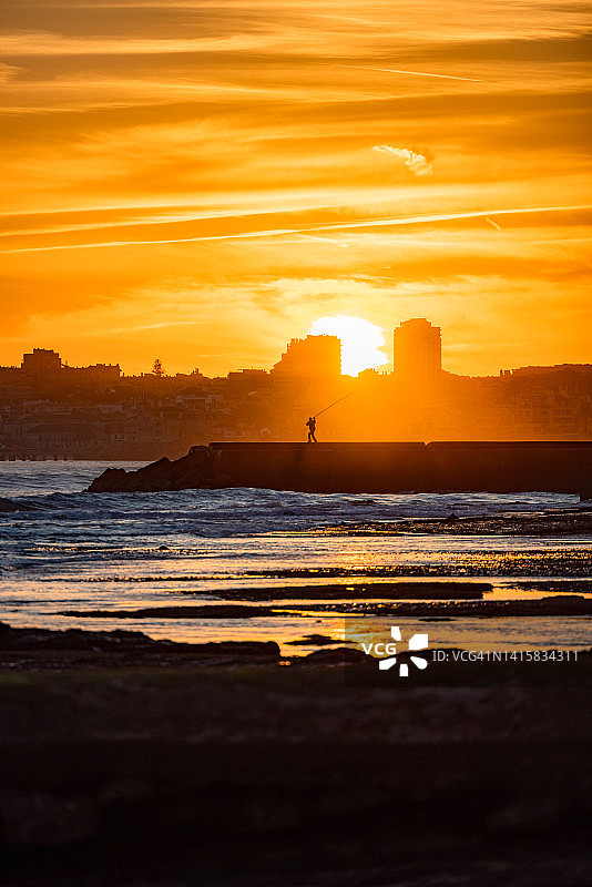 在码头钓鱼的男人，背景是戏剧性的日落和城市景观图片素材