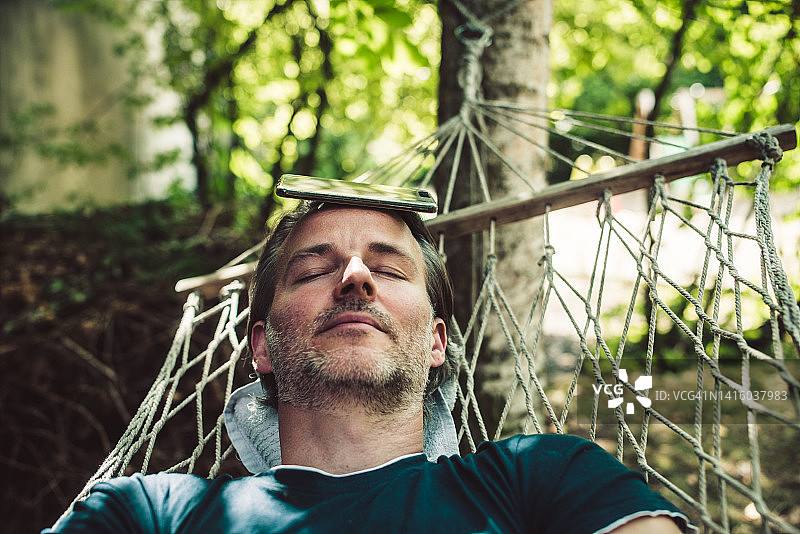 年长的男人睡在吊床上，手机放在头上图片素材