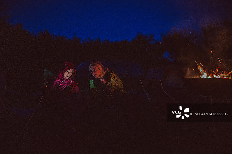 两个女朋友在露营地的篝火旁坐在露营椅上一起使用智能手机图片素材