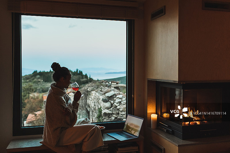 女人在壁炉旁放松，晚上喝红酒，用笔记本电脑看电影。图片素材