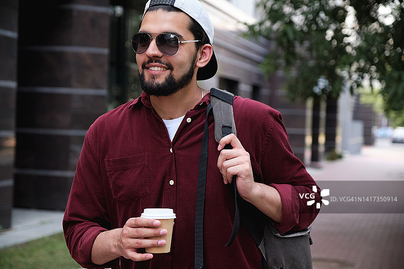 在城市中散步的微笑年轻男士的肖像。戴着棒球帽、喝着咖啡的深色皮肤印度男子。学生或游客在度假。交流生。徒步旅行。积极的情绪。图片素材