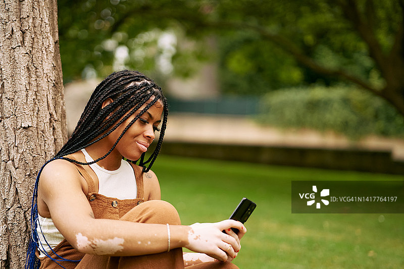 在公园休息时，一位患有白癜风的年轻女人正在用智能手机浏览社交媒体图片素材