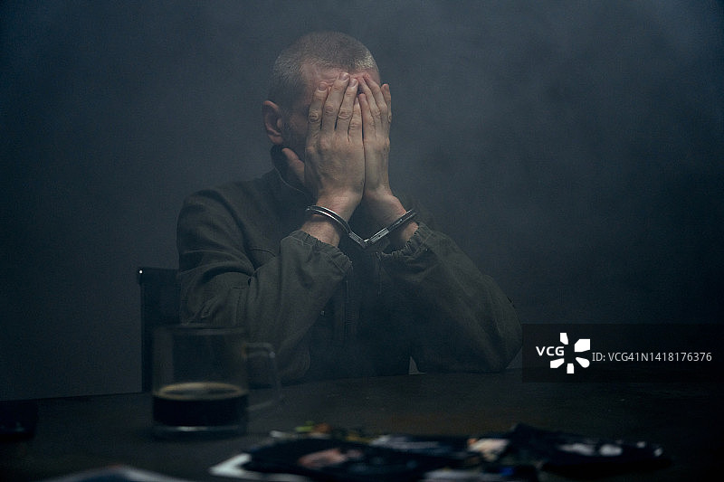 戴着手铐的罪犯坐在黑暗的雾气房间里，用手捂着脸图片素材